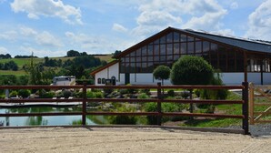 A large building with a sloped roof and large windows, surrounded by a fenced area with a pond and greenery.