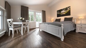 A bedroom with a large bed, a desk with chairs, a window with curtains, and a painting on the wall.