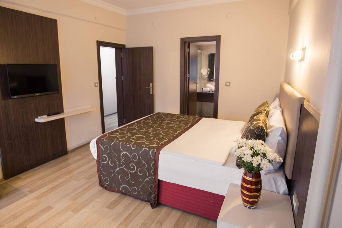 A hotel room with a bed, a TV mounted on the wall, a mirror, and a vase with flowers on the nightstand.