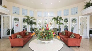A spacious lobby with two red sofas facing a central round table, surrounded by large windows and indoor plants. - Ov3718 - Windsor Palms Resort - 3 Bed 2 Baths Townhome (Kissimmee)