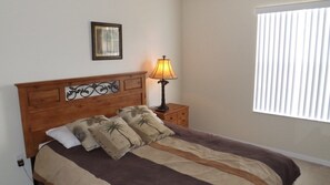 A neatly made bed with a striped bedspread and palm leaf pillows, a wooden headboard, a bedside lamp, a framed picture on the wall, and a window with blinds. - Ov3696 - Terra Verde Resort - 3 Bed 2.5 Baths Townhome (Kissimmee)