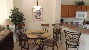 A dining area with a round glass table and six chairs, a lamp, a framed picture, and a kitchen in the background. - Ov3696 - Terra Verde Resort - 3 Bed 2.5 Baths Townhome (Kissimmee)