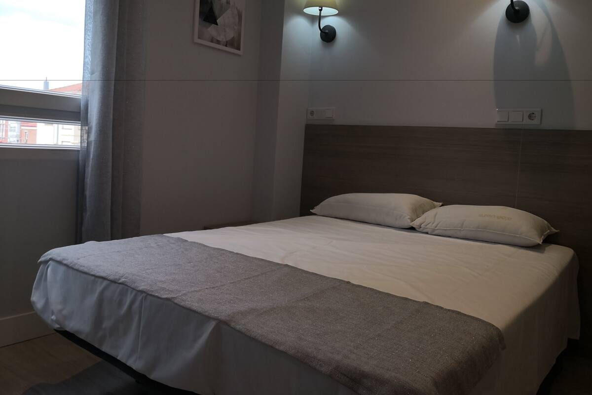 Uma cama bem arrumada com um cobertor cinzento e duas almofadas, um candeeiro fixado na parede e um quadro emoldurado.