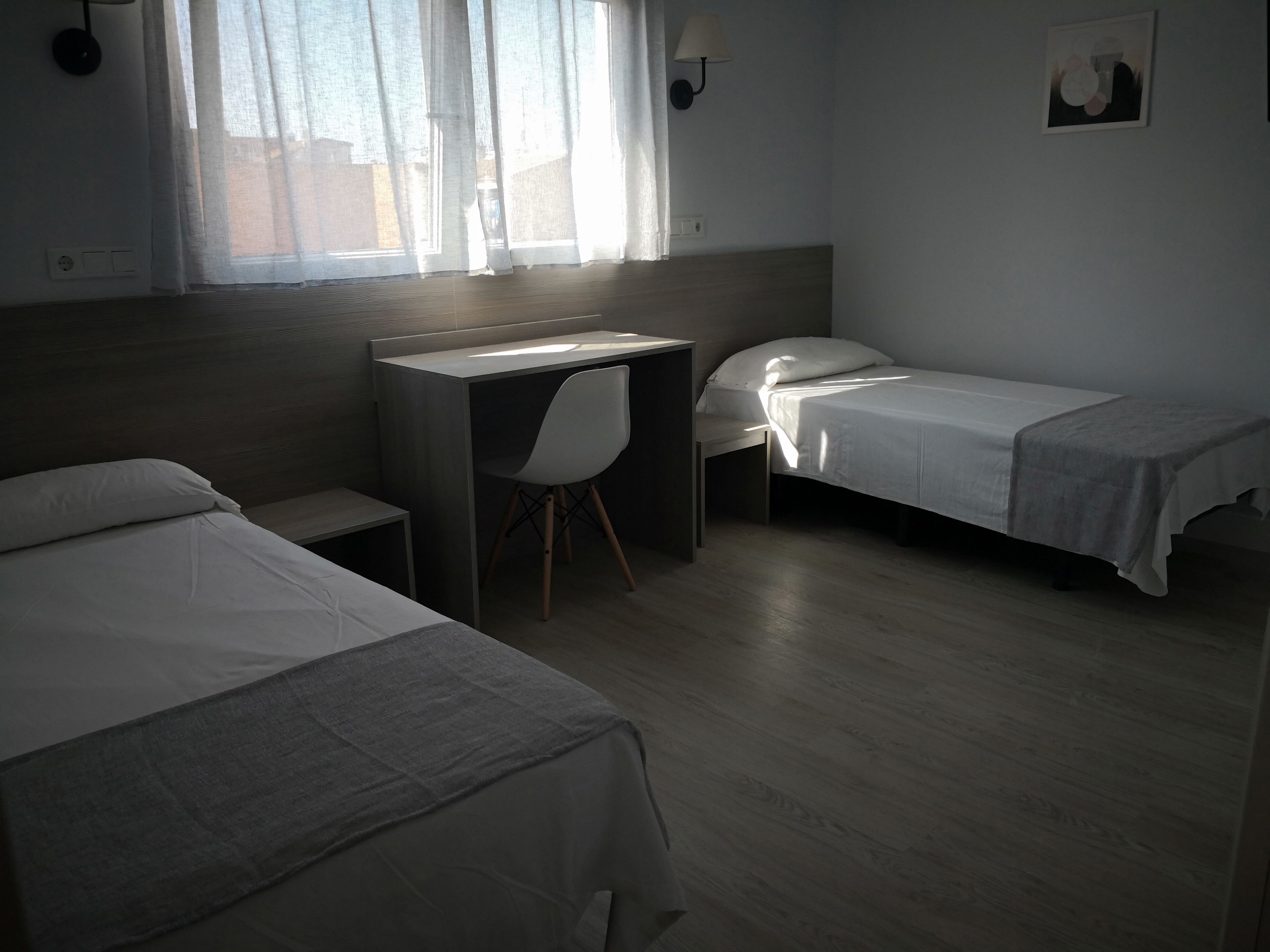 A hotel room with two single beds, a desk, a chair, and a window with sheer curtains.