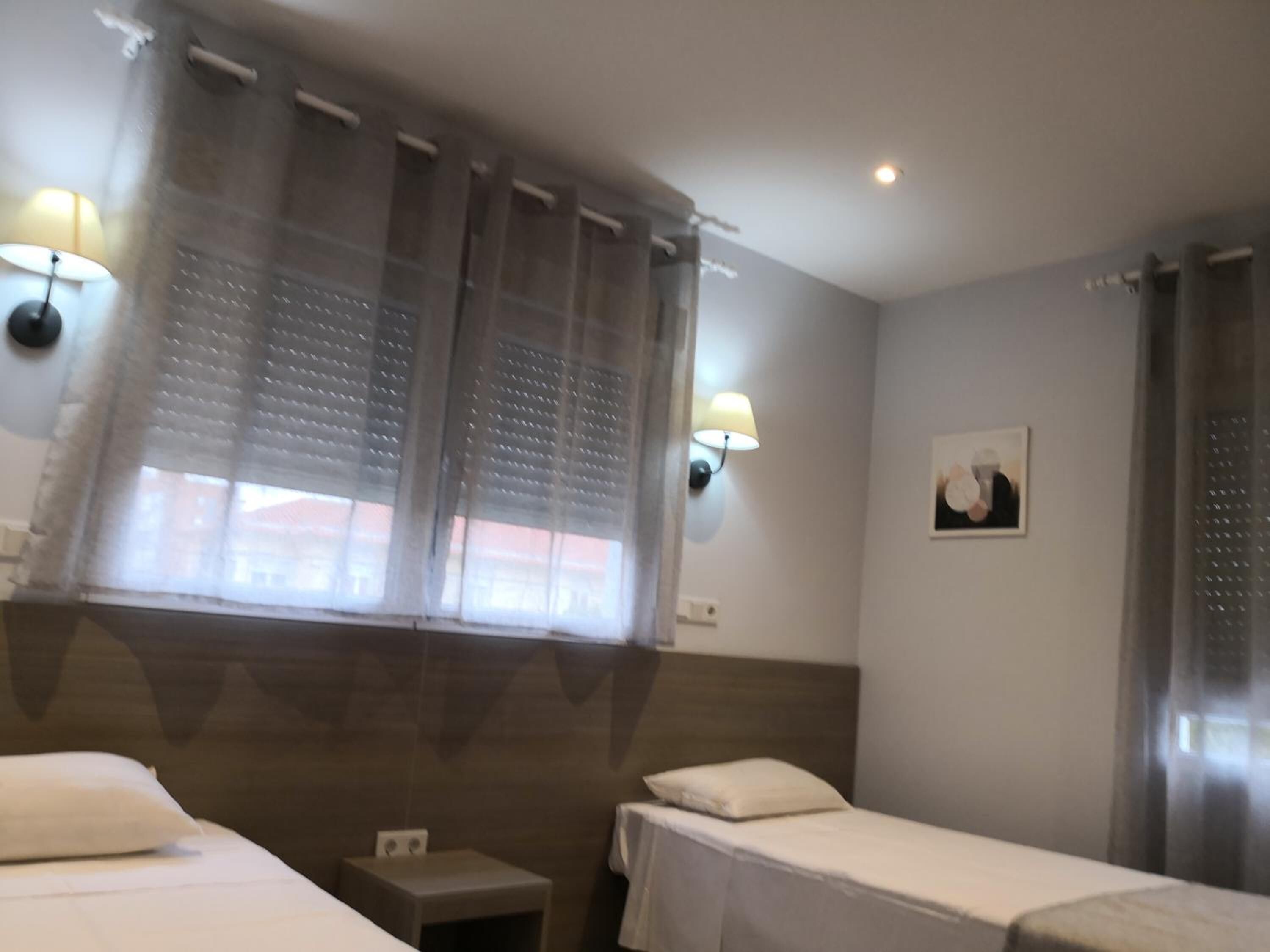 A hotel room with two single beds, a small bedside table, a wall-mounted lamp, and a window with sheer curtains.