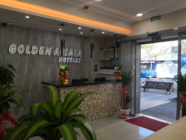 Zona d'entrada d'un hotel amb disseny modern, incloent un taulell de recepció, plantes en testos i un cartell que diu "GOLDEN ALCALA HOTELES".