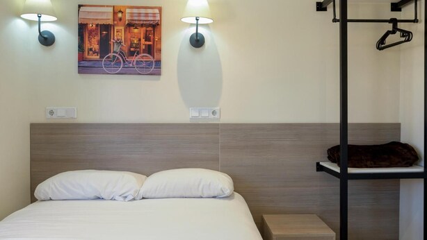 A neatly made bed with white linens, a headboard with a wood finish, two wall-mounted light fixtures, and a painting of a bicycle on the wall. - Hostal Golden Alcalá (Madrid)