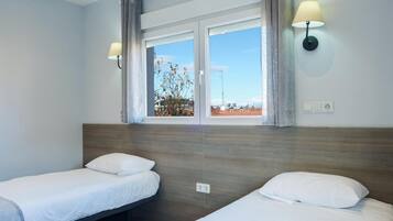 Habitación de hotel con dos camas individuales, un ventanal con cortinas y lámparas fijadas a la pared.