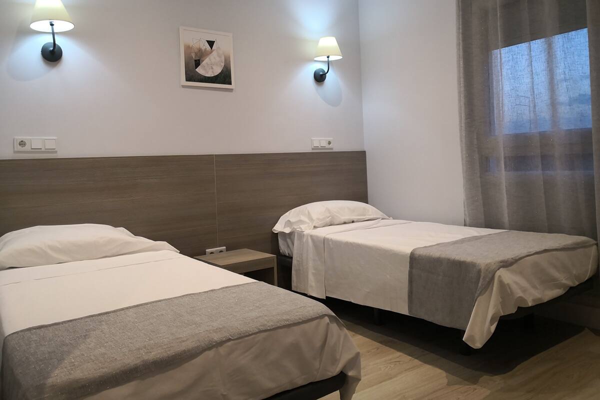 Quarto de hotel com duas camas, uma cabeceira de madeira, um quadro na parede e duas luminárias.