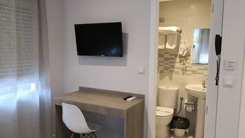Un baño moderno con inodoro, bidé y lavamanos. Cuenta con zona de ducha con puerta de cristal, y un pequeño escritorio con silla y televisor de pantalla plana montado sobre él.