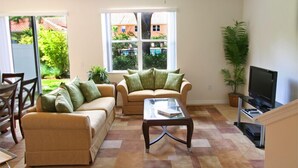 A living room with a sofa, a coffee table, and a television. There is a window with a view of a garden and a building.