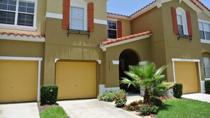 A two-story house with a yellow facade, red shutters, and a small front yard with a palm tree and shrubs.