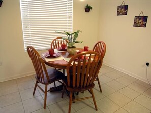 Een eetgedeelte met een ronde tafel, vier houten stoelen, een potplant en twee aan de muur hangende schilderijen.