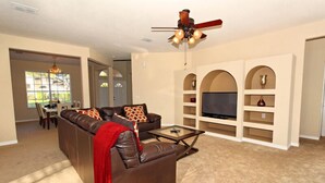 A living room with a brown leather sofa, a glass coffee table, a ceiling fan, and built-in shelving with a flat-screen TV.