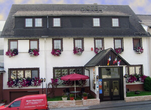 A hotel with a white facade, multiple windows with flower boxes, a red van parked in front, and a small outdoor seating area with a red umbrella.