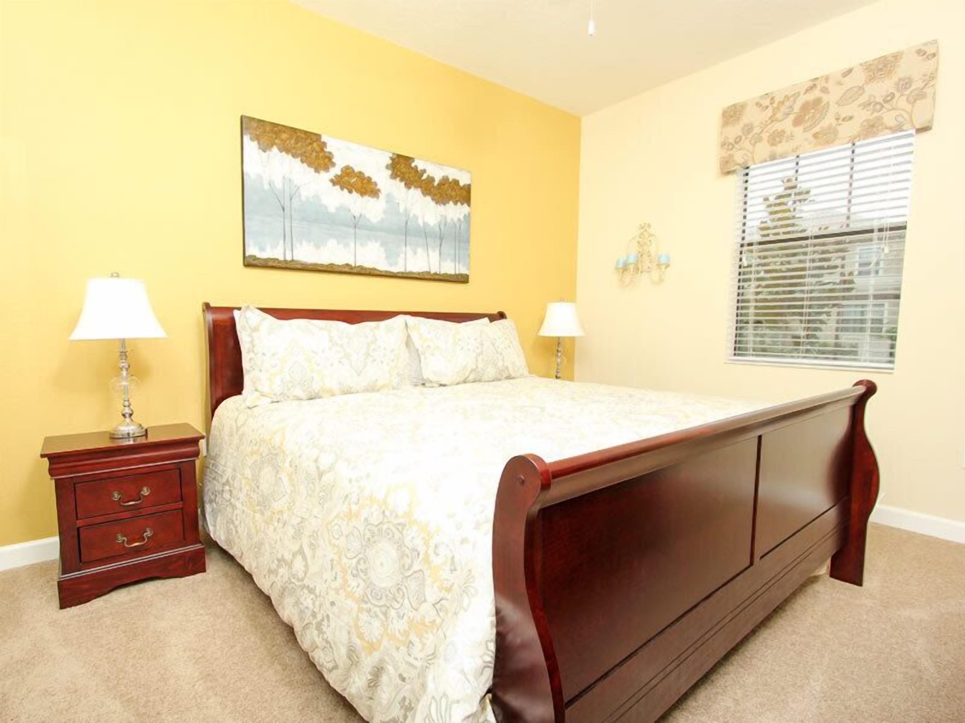 A bedroom with a wooden bed, two bedside tables, a nightstand, a painting on the wall, and a window with blinds.