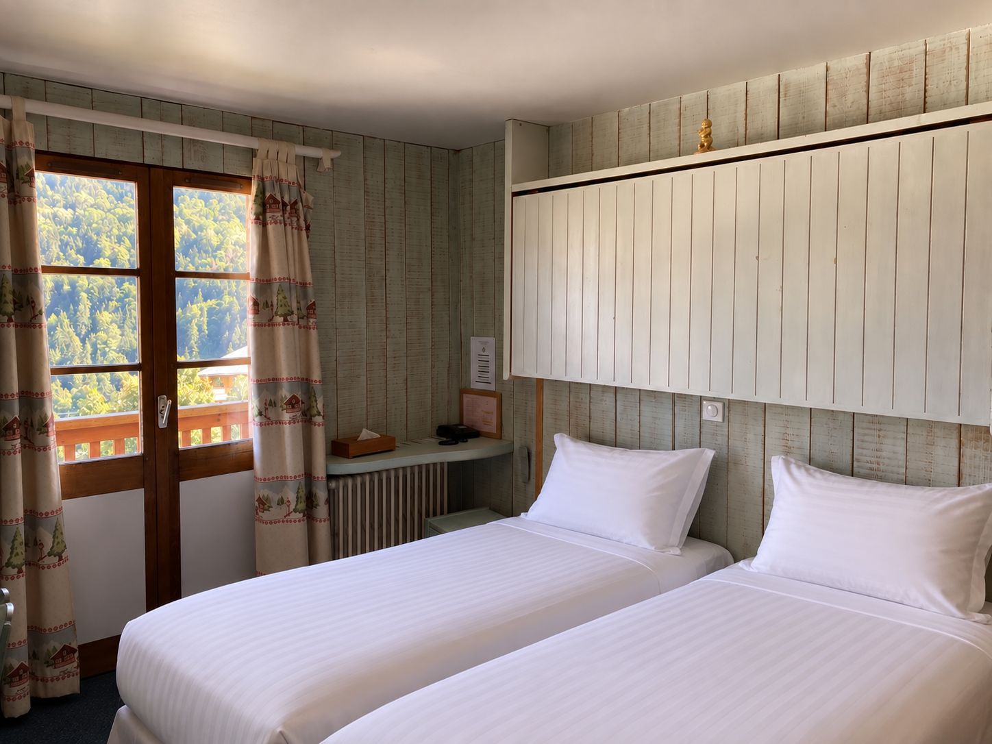 A room with two beds, a wooden headboard, a window with curtains, and a small table with a tissue box.