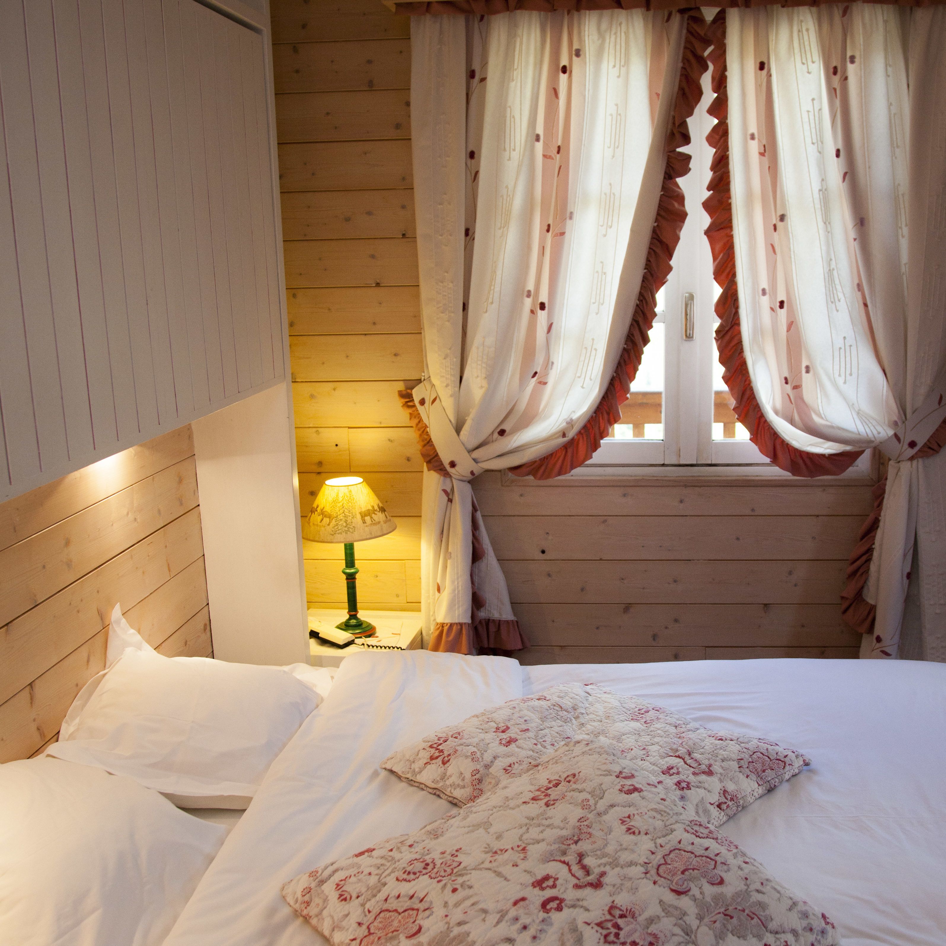 Ein Schlafzimmer mit hölzernem Kopfteil, einem Bett mit weißen Leinentüchern und Kissen mit Blumenmuster, einem Nachttisch mit Lampe sowie einem Fenster mit Vorhängen.