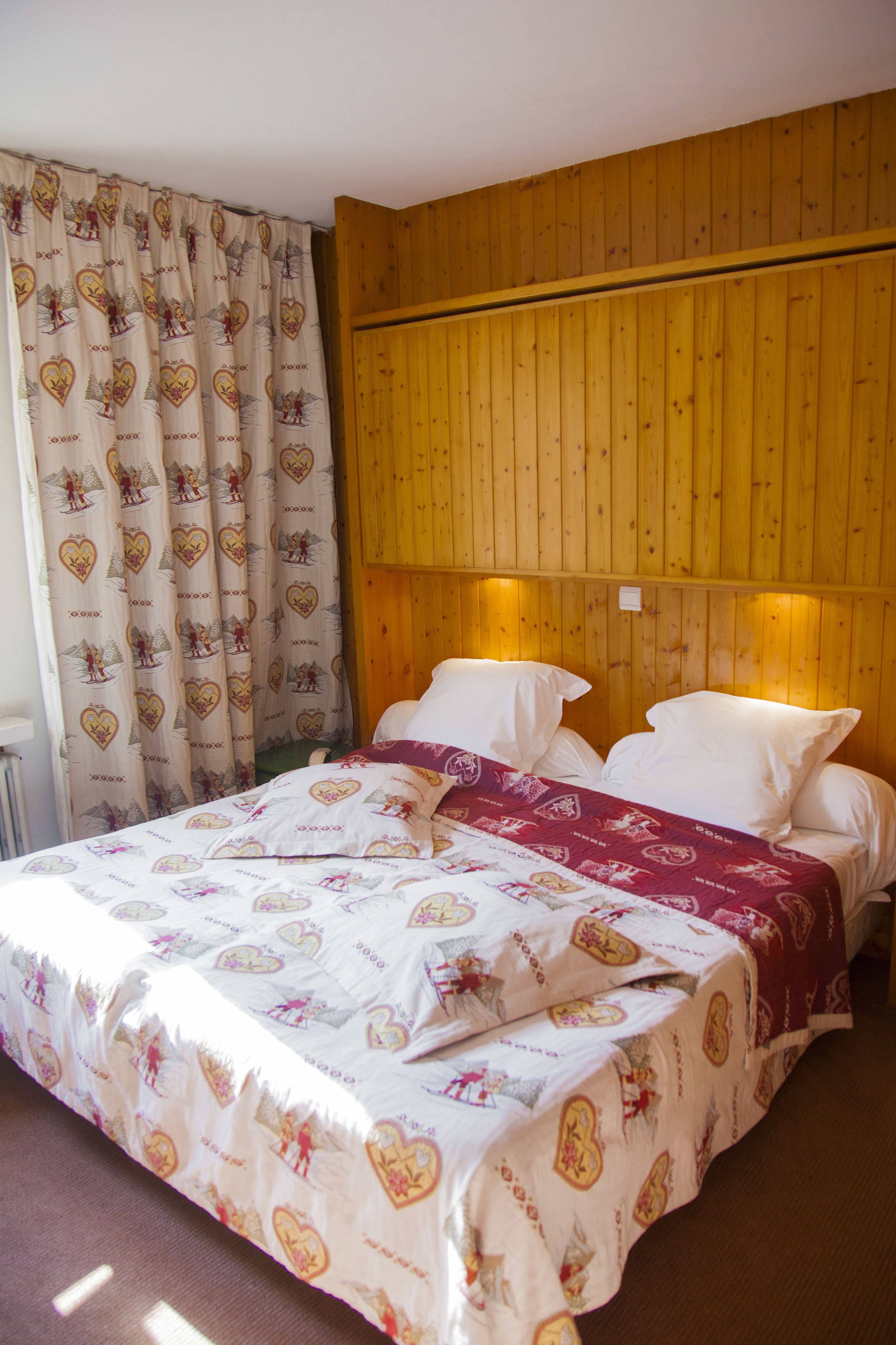 Une chambre à coucher avec une tête de lit en bois, un lit avec une literie blanche et rouge, et un rideau orné d’un motif en forme de cœur.