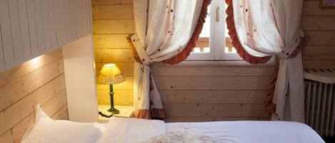 A bedroom with a wooden headboard, a bed with white linens and floral-patterned pillows, a nightstand with a lamp, and a window with curtains.