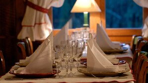 Une table à manger dressée avec des serviettes blanches, des verres à vin et des assiettes, une lampe et une fenêtre en arrière-plan.