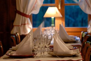 Una mesa de comedor dispuesta con servilletas blancas, copas de vino y platos, con una lámpara y una ventana al fondo.