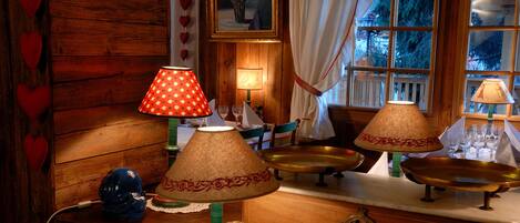 A wooden dining room with a table set for four, featuring a chandelier, red and white heart decorations, and a window with curtains.