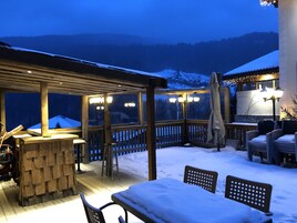 Un patio exterior cubierto con mobiliario de madera y una barra, rodeado de nieve y montañas al fondo.