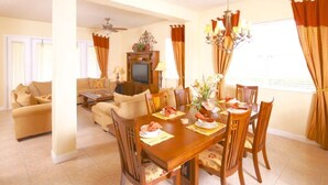 Un coin repas avec une table en bois dressée pour quatre personnes, entourée de chaises. On aperçoit un téléviseur au fond, un ventilateur de plafond et un lustre au-dessus de la table.