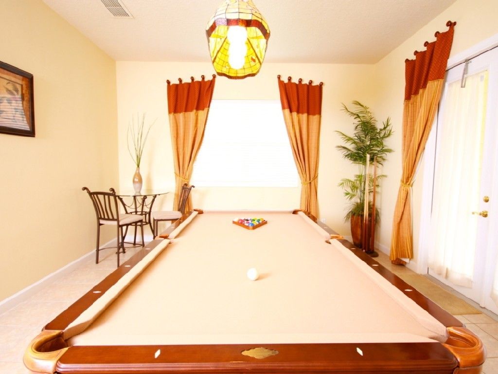 Une table de billard recouverte d’un drap blanc, entourée de rideaux orange, d’une lampe suspendue et d’une petite table avec des chaises.