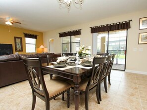 A dining area with a table set for four, surrounded by chairs, in a room with a ceiling fan, a sofa, and a large window with blinds.