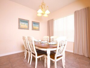 A dining area with a round table, six chairs, two framed pictures on the wall, a chandelier, and a window with curtains.