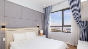 Una habitación de hotel moderna con una cama grande, una mesita de noche, una lámpara y vistas a un paisaje urbano a través de un ventanal amplio.
