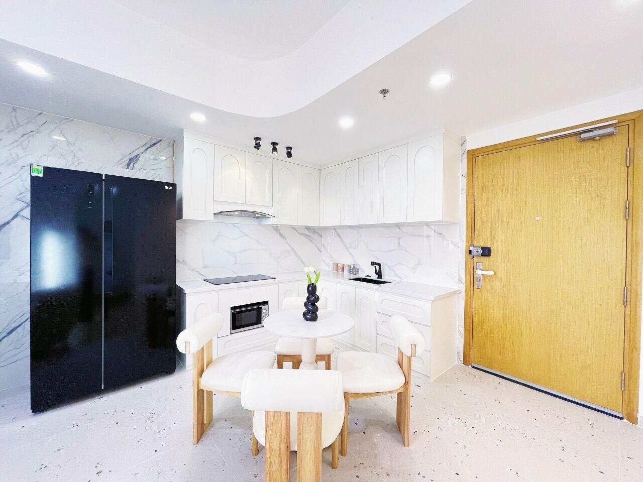 A modern kitchen with white cabinetry, a black refrigerator, a wooden dining table with white chairs, and a wooden door.