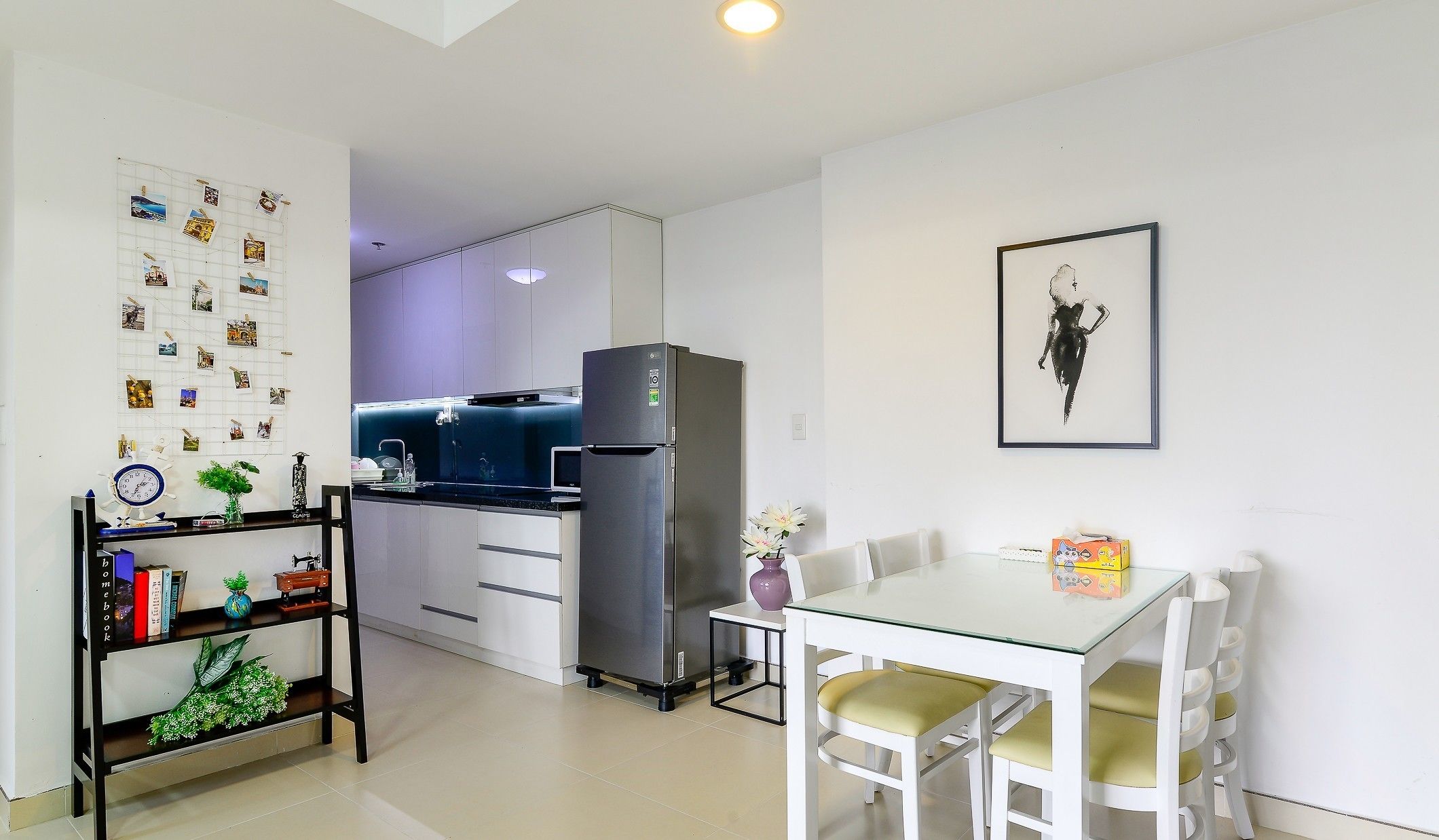 Una cocina compacta con refrigerador, una mesa de comedor con sillas, un estante pequeño con libros y un reloj, y una pared decorada con fotografías.