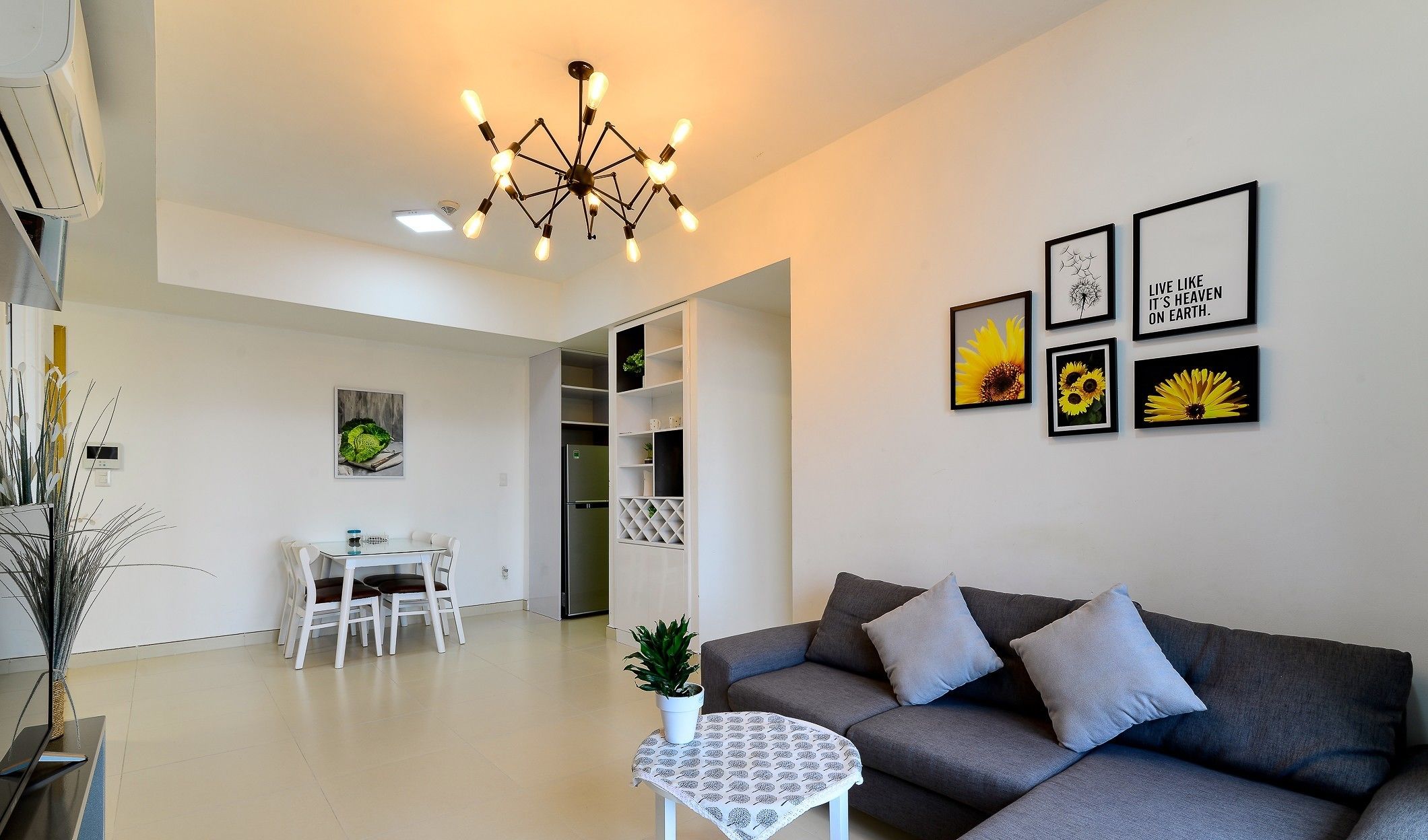 A modern living room with a grey sofa, a small round table, and a dining area with a white table and chairs. There are framed pictures on the wall and a unique light fixture above the sofa.
