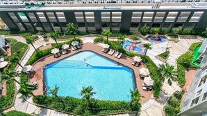 Una piscina grande y rectangular rodeada de sillas reclinables y palmeras, con un área de juegos cerca.