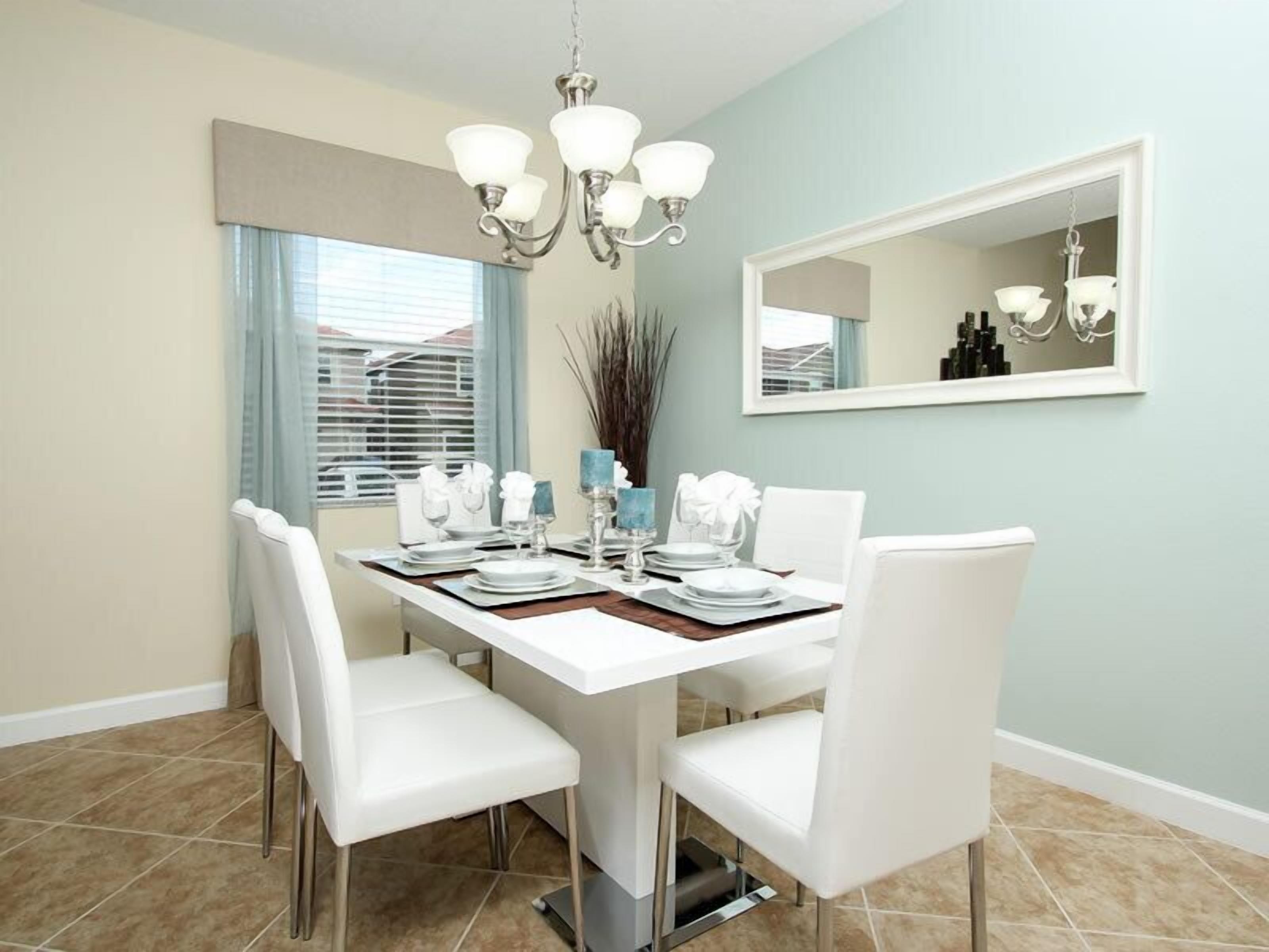 A dining area with a white table set for four, surrounded by white chairs. A chandelier hangs above the table, and a mirror is mounted on the wall to the right.