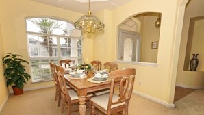 A dining room with a wooden table set for four, surrounded by chairs, a chandelier, and a large window with blinds.