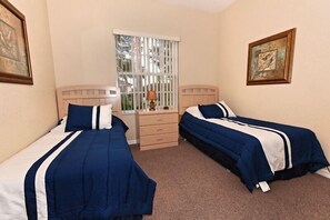 A room with two beds, a window with blinds, a bedside table with a lamp, and framed artwork on the walls.