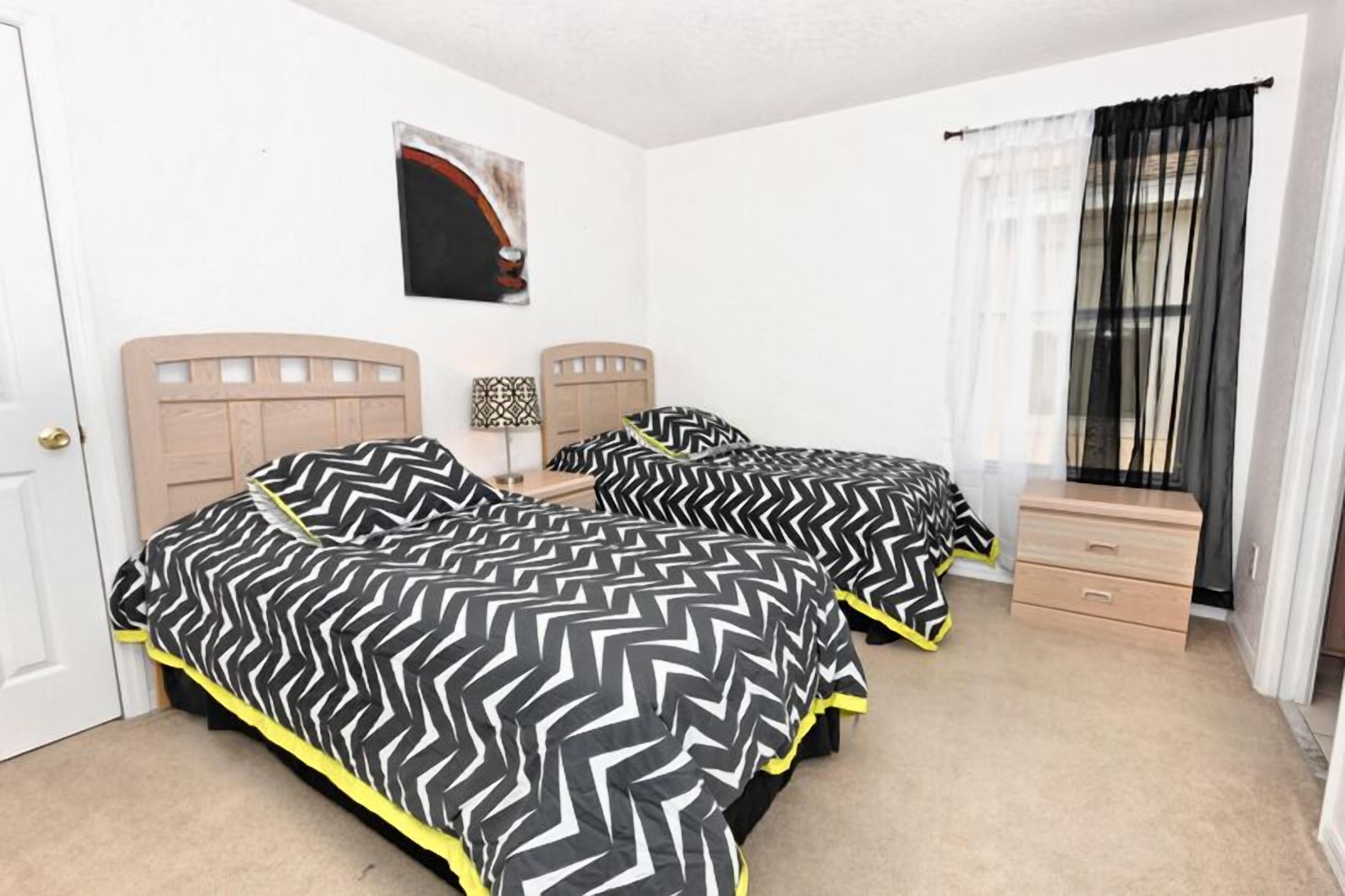 Une chambre avec deux lits, une table de chevet, une commode et une fenêtre avec des rideaux légers.