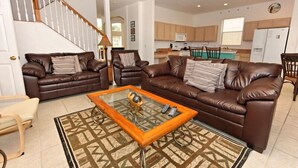 A spacious living room with two brown leather sofas, a glass coffee table, and a patterned rug. A staircase is visible in the background leading to an upper floor.