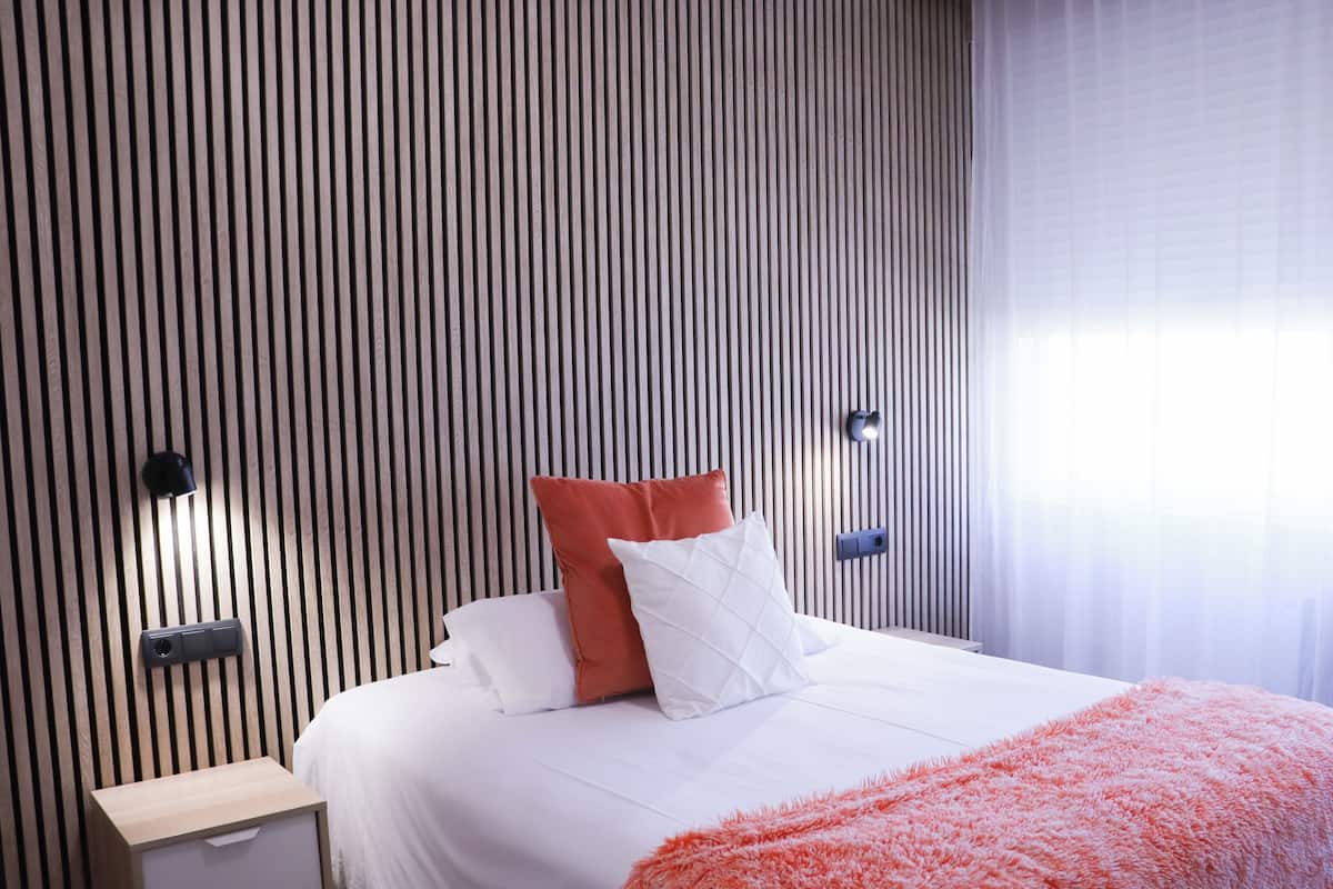 A hotel room with a bed, a nightstand, a wall-mounted lamp, and a window with curtains.