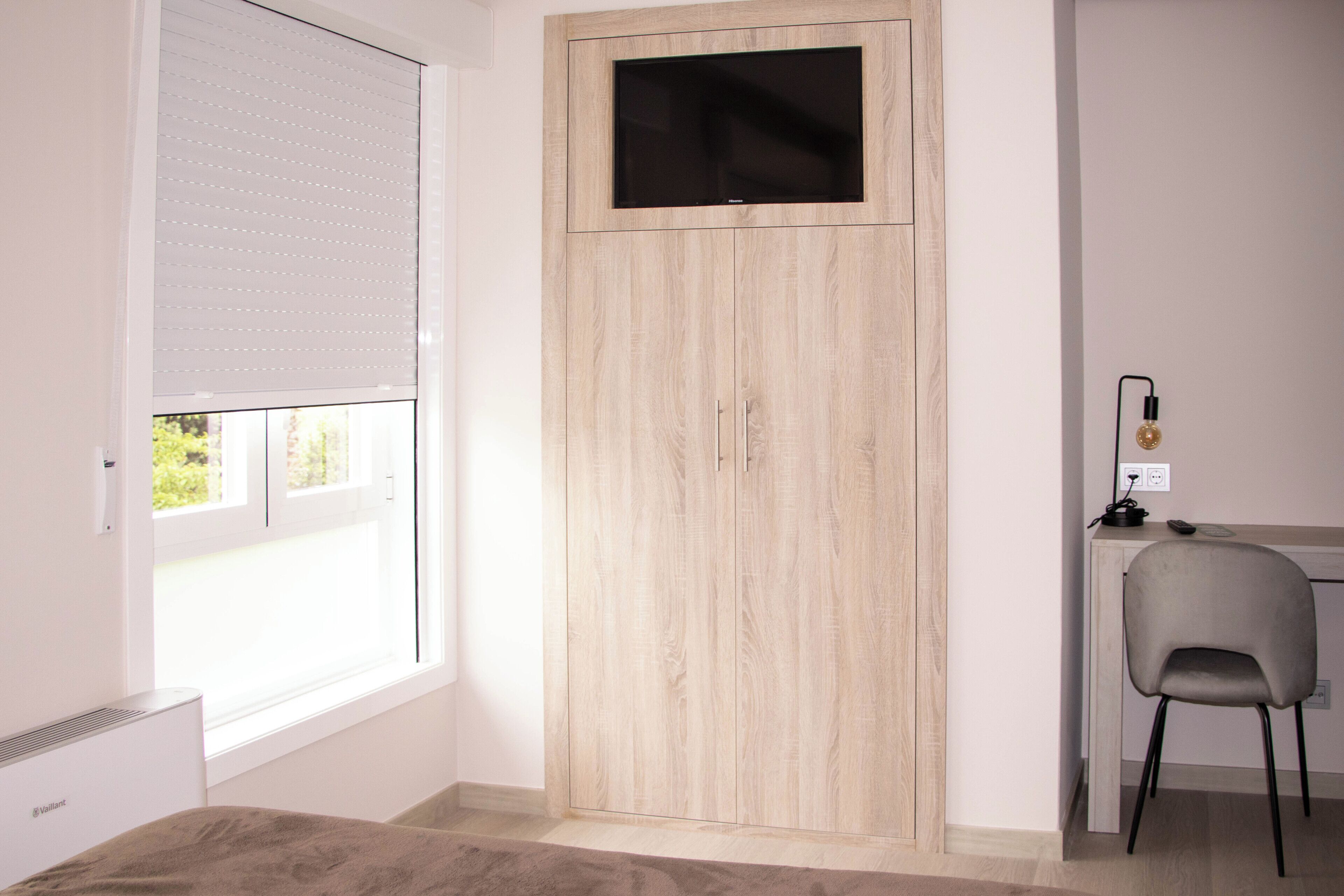 Een kamer met een kast, een bureau met stoel, een raam met zonwering en een tv aan de muur.