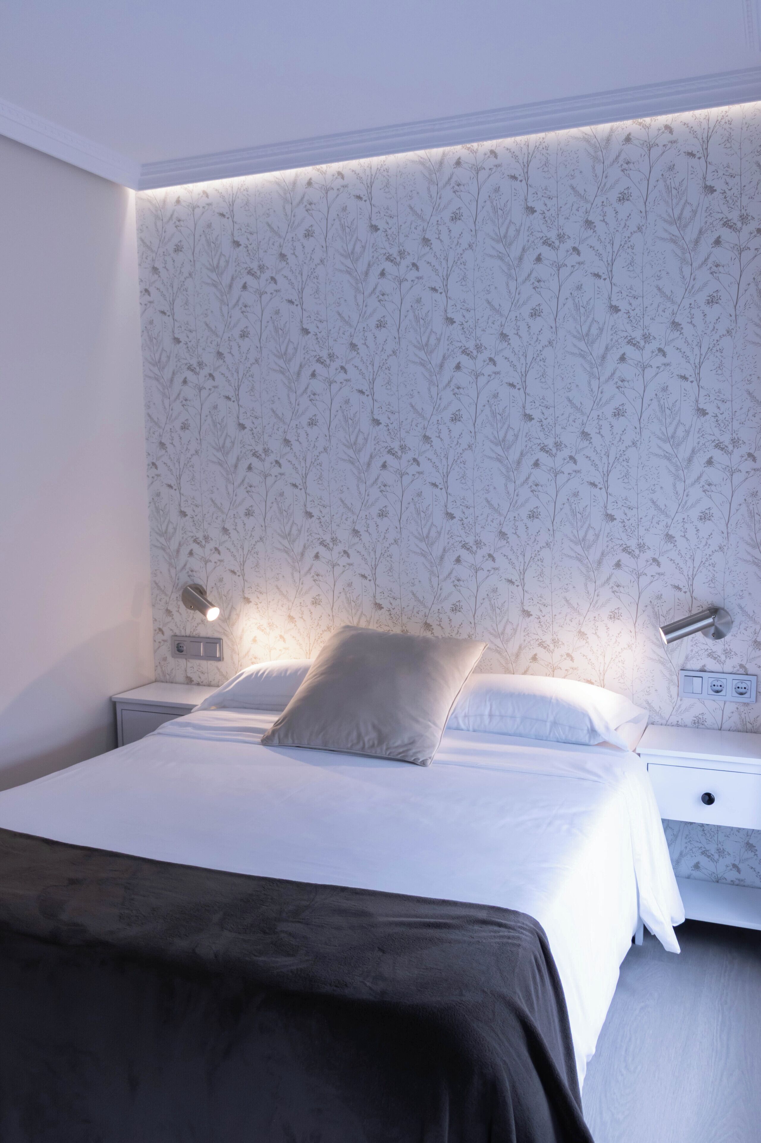 A modern bedroom with a patterned wallpaper, a bed with white linens, two bedside lamps, and a nightstand with a power outlet.