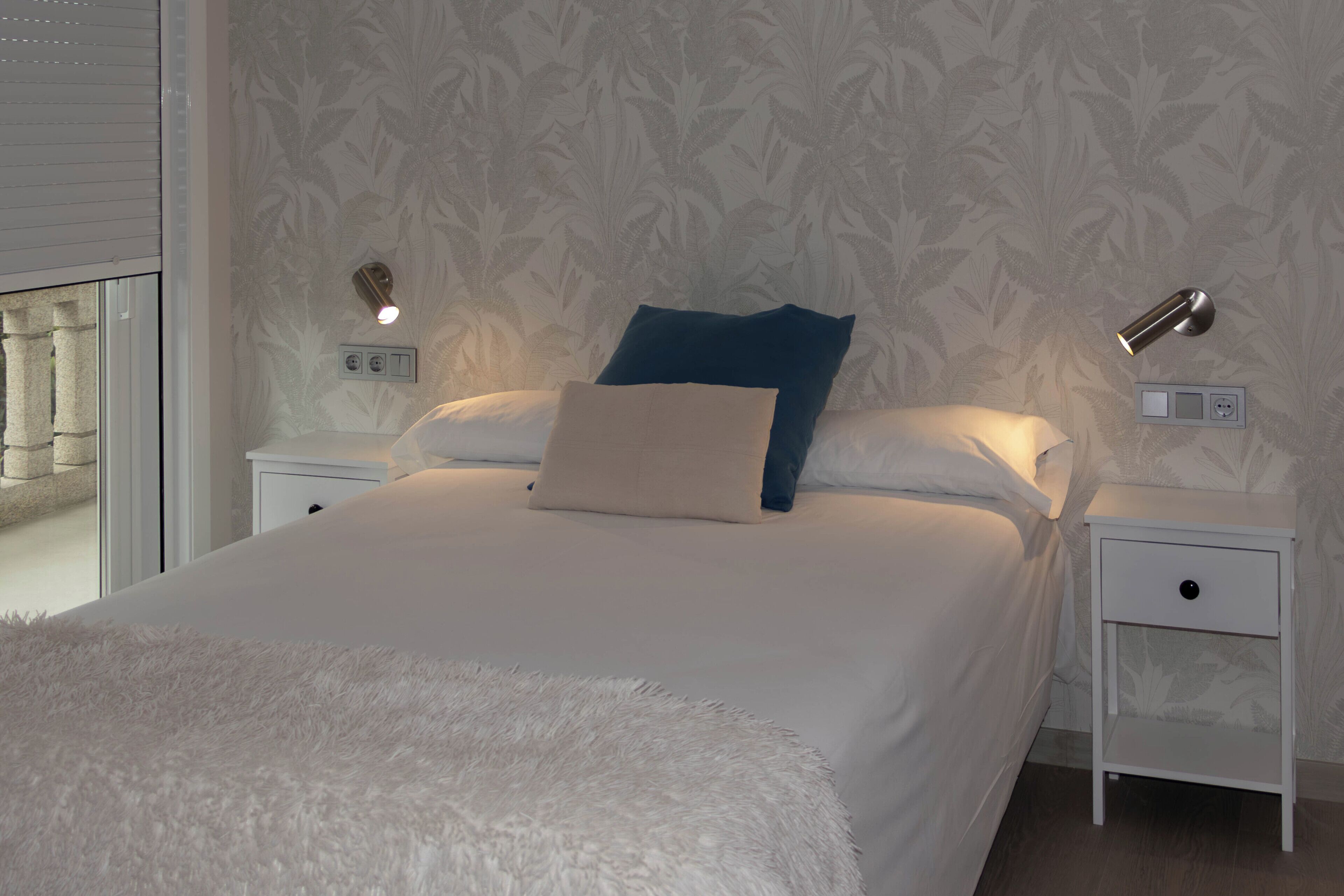 A neatly made bed with a fluffy white cover, two bedside tables with lamps, and a patterned wallpaper.