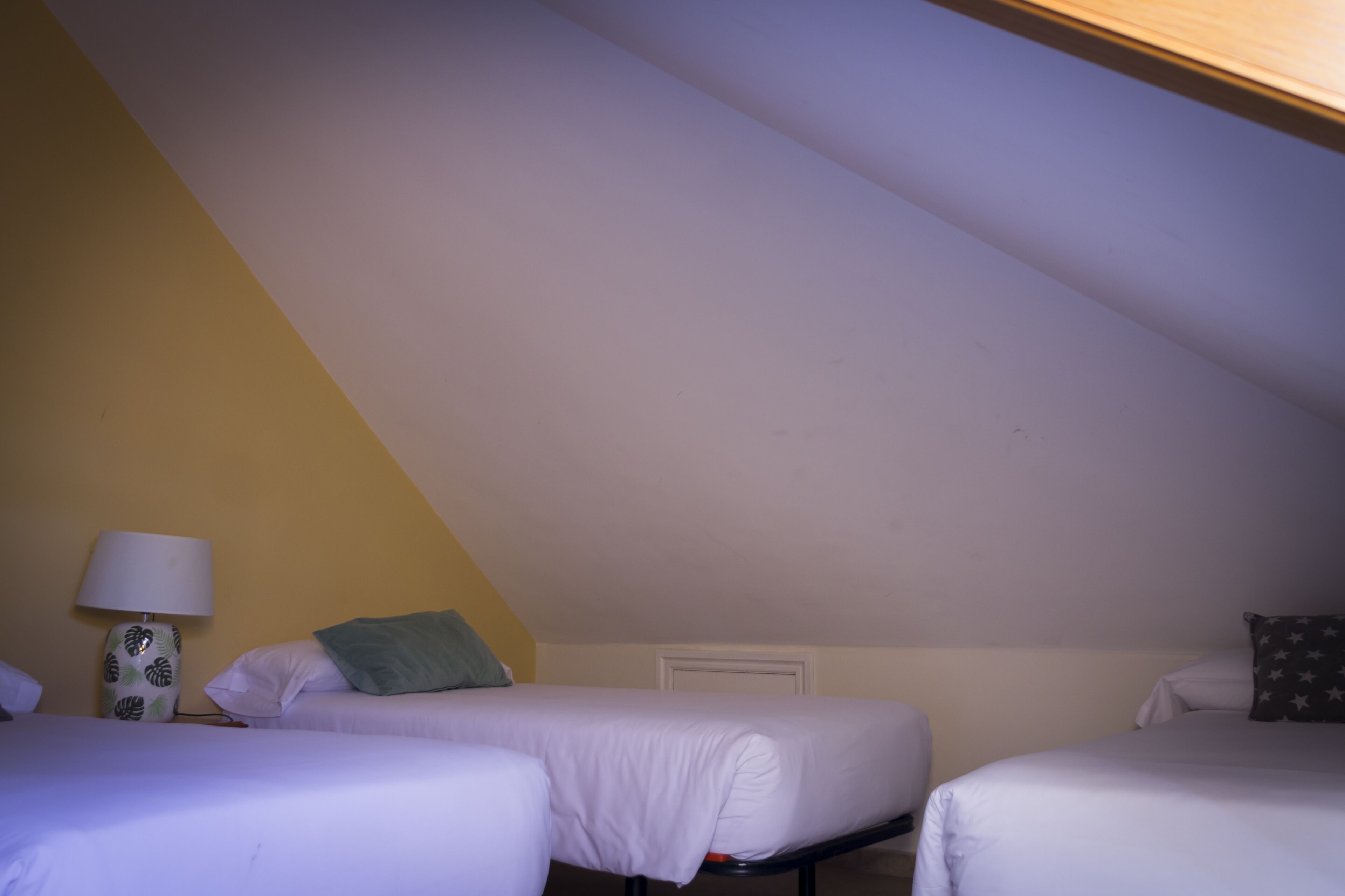 Een kleine, zwak verlichte kamer met twee eenpersoonsbedden, een nachtkastje met een lamp en een eenvoudige muur.