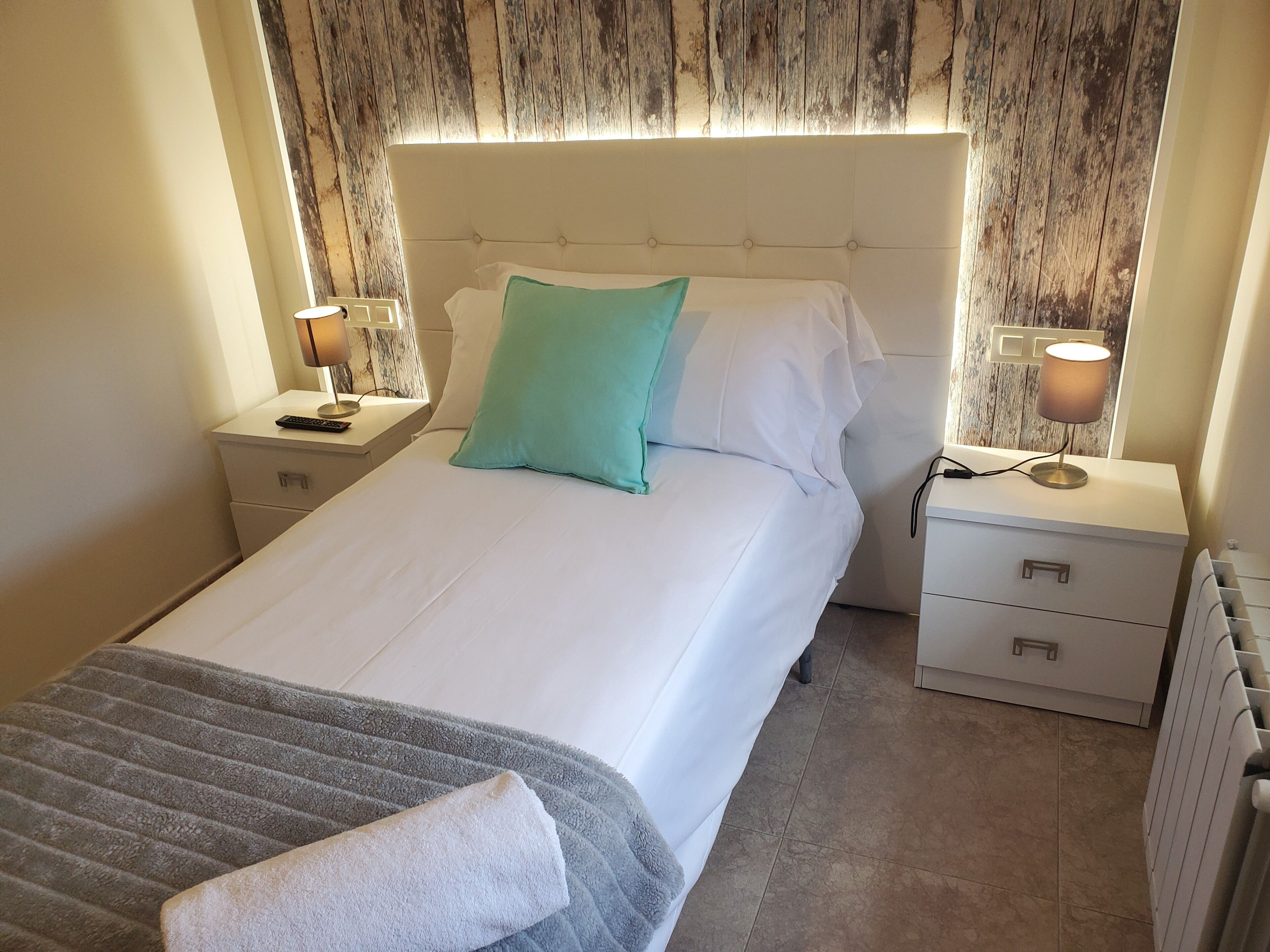 Una cama bien tendida con una colcha blanca y almohadones verde azulado, con mesitas de noche con lámparas y un tocador con un control remoto.