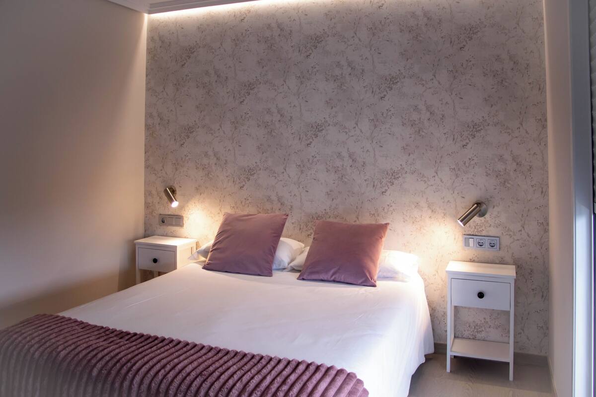 A neatly made bed with two pillows, a bedside table with a lamp, and a wall-mounted switch.