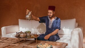 A person pouring tea from a traditional teapot into a cup, seated on a white sofa with a tray of teaware and a plate of cookies in front. - Tagadert Lodge (Tameslouht)